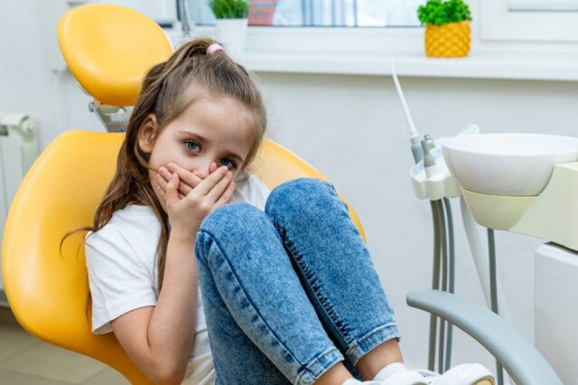 Young girl sitting in a dental chair, covering her mouth with her hands, displaying signs of dental anxiety in a bright dental office setting.