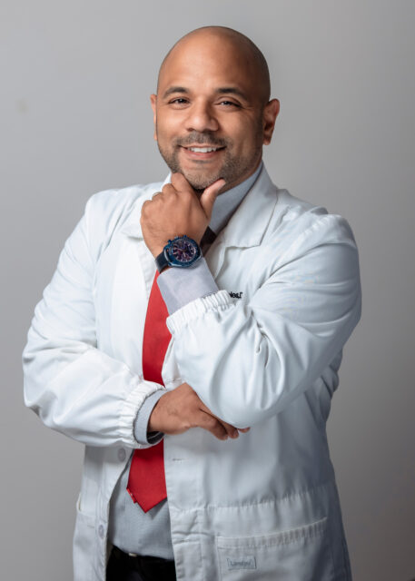 Dr. Kenny Alejandro Robles in a white dental coat with a red tie, smiling confidently and posing with his hand on his chin, representing his commitment to quality dental care at Robles Family Dental.