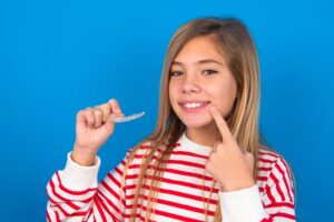 Girl smiling and holding clear Invisalign aligner, demonstrating confidence in orthodontic treatment against a blue background.